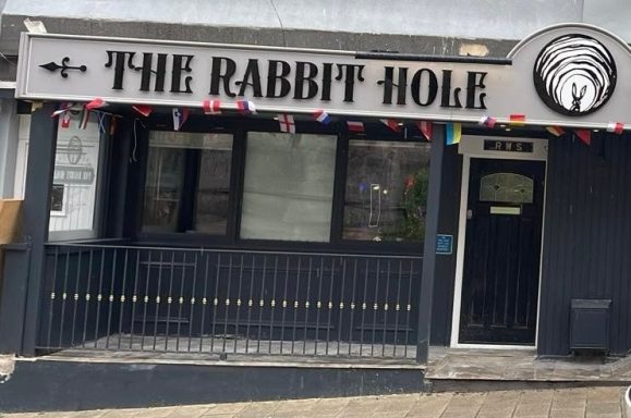 Front view of "The Rabbit Hole" establishment with a decorative sign and outdoor seating.