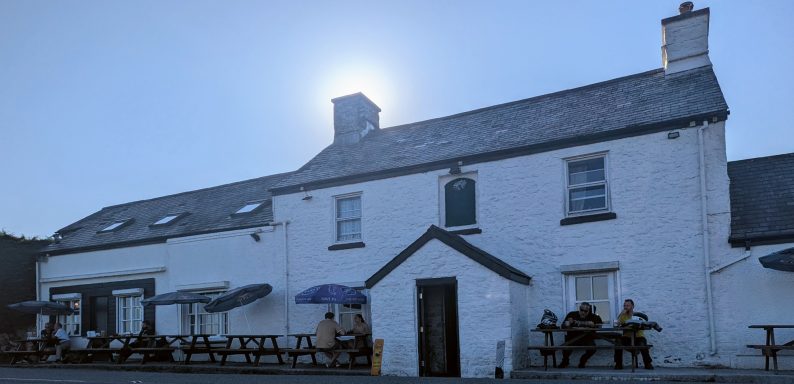 A white building with a slate roof, outdoor seating, and a sun shining above.