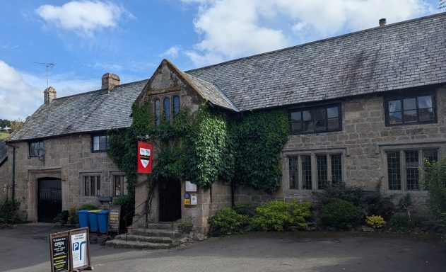 Historic stone building with a slate roof, surrounded by greenery and a blue sky.