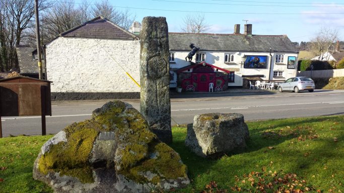 Stone monolith and surrounding rocks in a rural setting with a building and vehicles in the background.