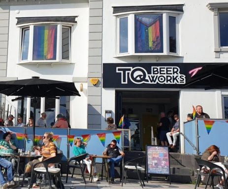Lively outdoor area of a bar decorated with rainbow flags and patrons seated at tables.