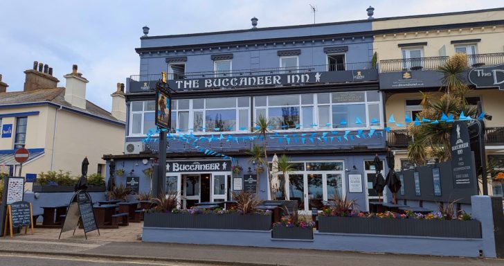 A two-storey blue-painted pub with outdoor seating and surrounding plants.