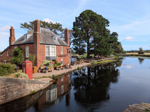 Quaint house by a calm river, surrounded by greenery and a red phone box nearby.
