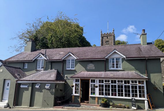 Green cottage with a steep roof, white trim and a church tower in the background under a blue sky.
