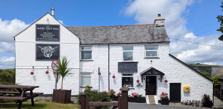 A white building with a sign, surrounded by plants, under a blue sky with clouds.