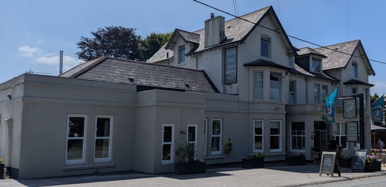 A two-storey building with a grey exterior and pitched roofs, surrounded by trees.