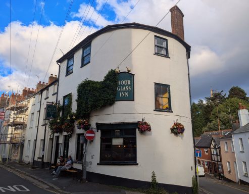 A quaint, curved white building with green ivy, featuring hanging flower baskets and a sign.