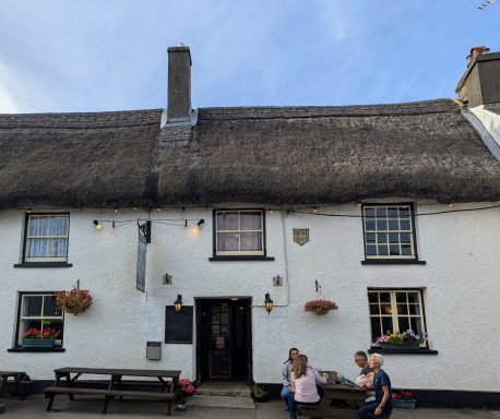 Thatched-roof pub with flower baskets and outdoor seating, two people walking nearby.