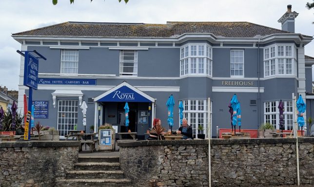 Grey-painted pub with outdoor seating, colourful flags, and patrons enjoying the atmosphere.