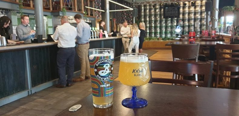 Two glasses of beer sit on a table in a busy bar with customers in the background.
