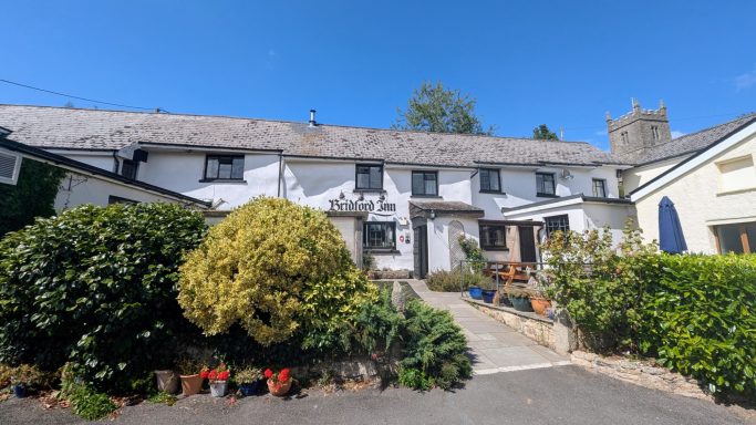 White cottage with a thatched roof, surrounded by greenery and flowers under a clear blue sky.