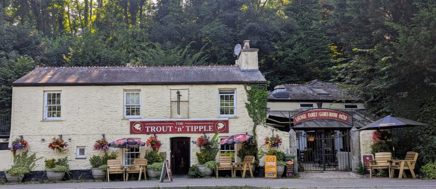 A quaint pub exterior surrounded by greenery, with outdoor seating and flower arrangements.