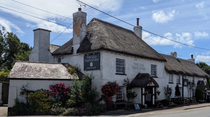 Thatched cottage with flower beds and a sign, under a blue sky with clouds.