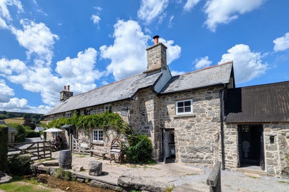 Stone cottage with a slate roof and chimney, surrounded by blue skies and fluffy clouds.