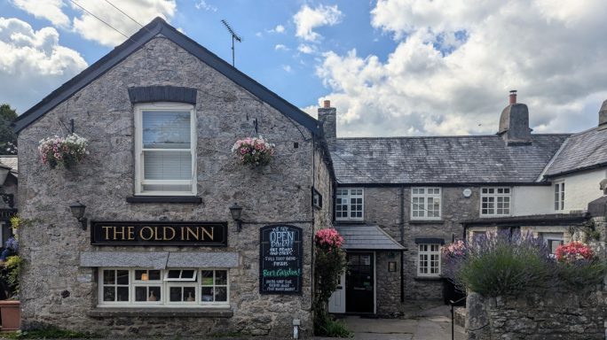 Stone building with a sign for "The Old Inn" and floral decorations under a cloudy sky.