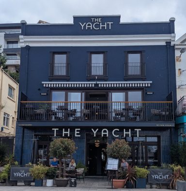Exterior of a blue pub named "The Yacht" with outdoor seating and plants.