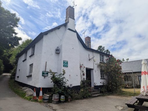 White cottage with a sloping roof, surrounded by greenery and gravel path.